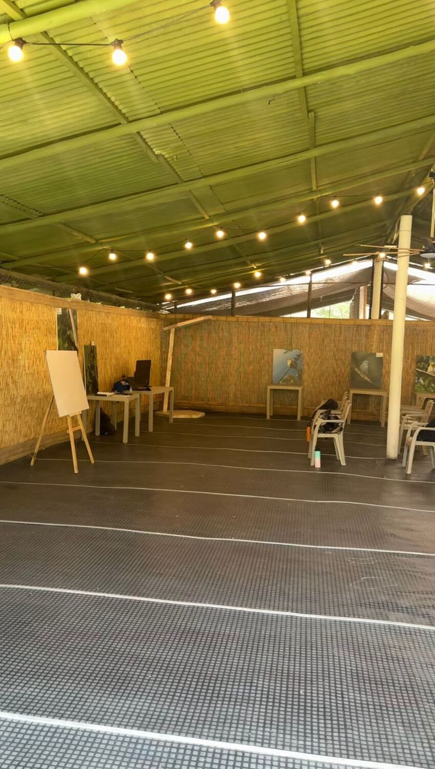 Taller techado: cubierta de caña, paredes en estera, guirnaldas de bombillos, un caballete y sillas Rimax blancas.