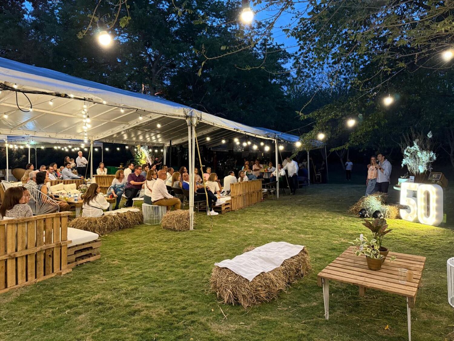 Fiesta de cumpleaños número 50 al anochecer: invitados conversando bajo una carpa blanca con bombillas, salas de palés, pacas de heno y letras luminosas con el número 50 sobre el prado.