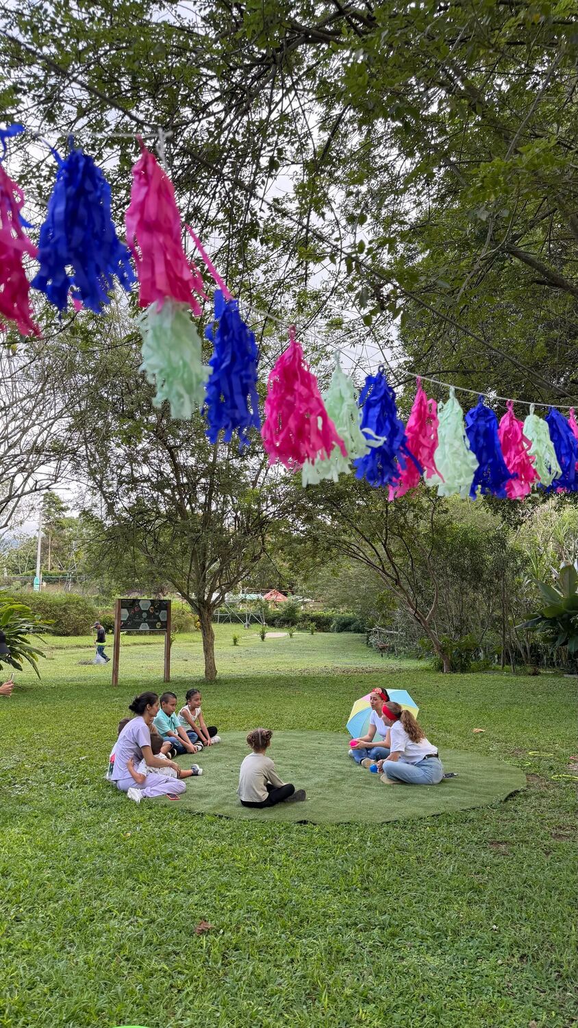 Niñas y niños sentados en círculo sobre el césped junto a dos recreacionistas con sombrilla, bajo una larga guirnalda de flecos azules, rosados y verdes colgada entre los árboles.