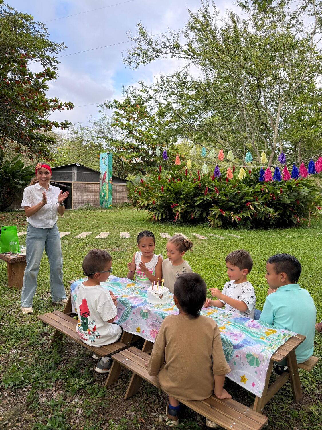 Seis niños alrededor de una torta con velas encendidas en una mesa de picnic de madera, mientras una recreacionista aplaude; al fondo, arbustos florecidos y guirnaldas de flecos.