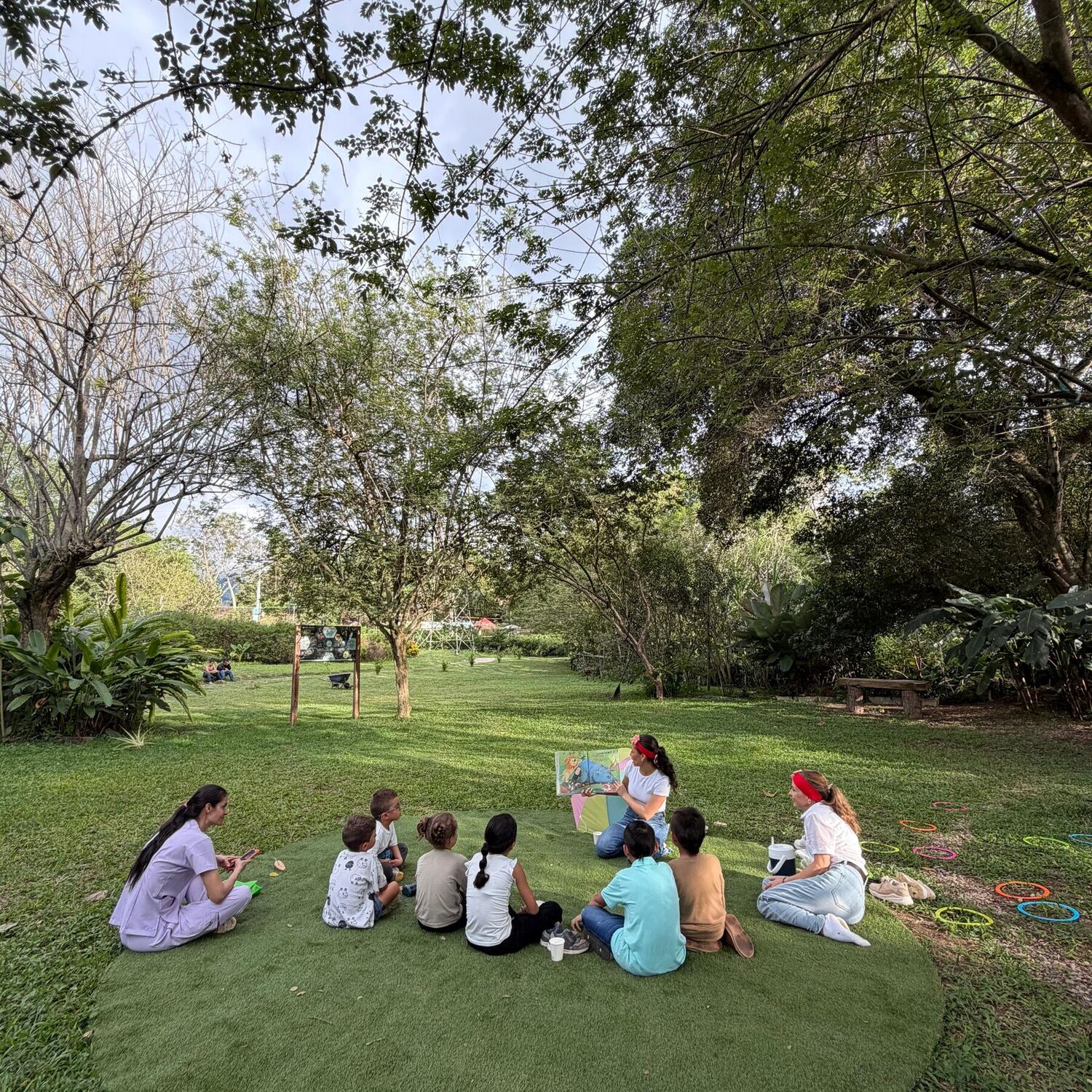 Cuentos al parque: dos recreacionistas leen un libro ilustrado a ocho niños sentados en círculo sobre una alfombra de césped, rodeados de árboles y aros de colores.