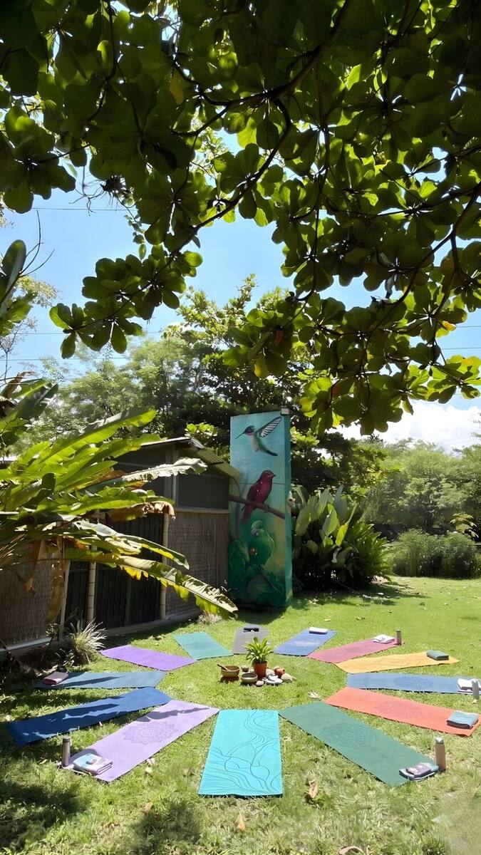 Doce colchonetas de colores dispuestas en círculo como los pétalos de una flor sobre el césped, con un centro de plantas y velas, frente al mural de aves y bajo la sombra de un árbol.