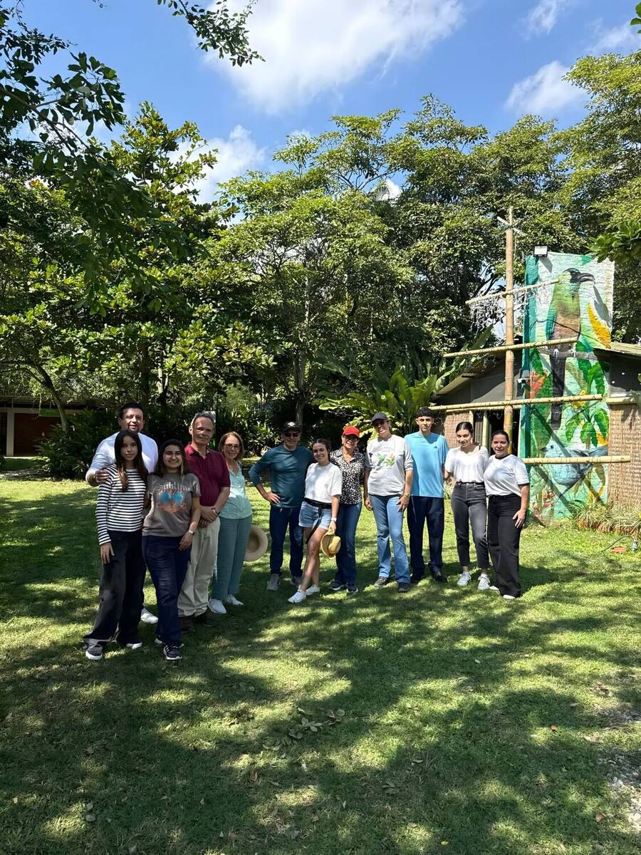 Grupo de trece personas posando de pie sobre el césped, a la sombra de los árboles y junto al mural pintado con aves del parque.
