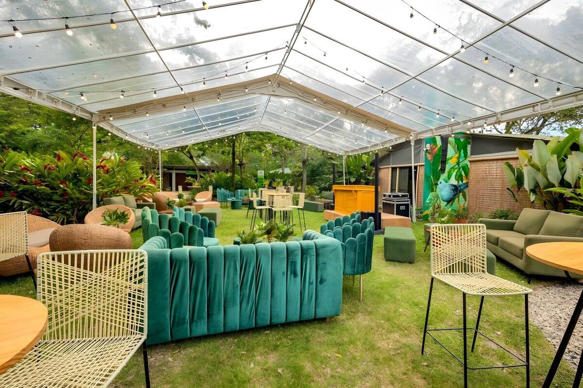 Montaje corporativo sobre el césped bajo una carpa transparente con luces colgantes: sofás verdes, poltronas de mimbre, mesas altas y un bar amarillo junto al mural de aves.