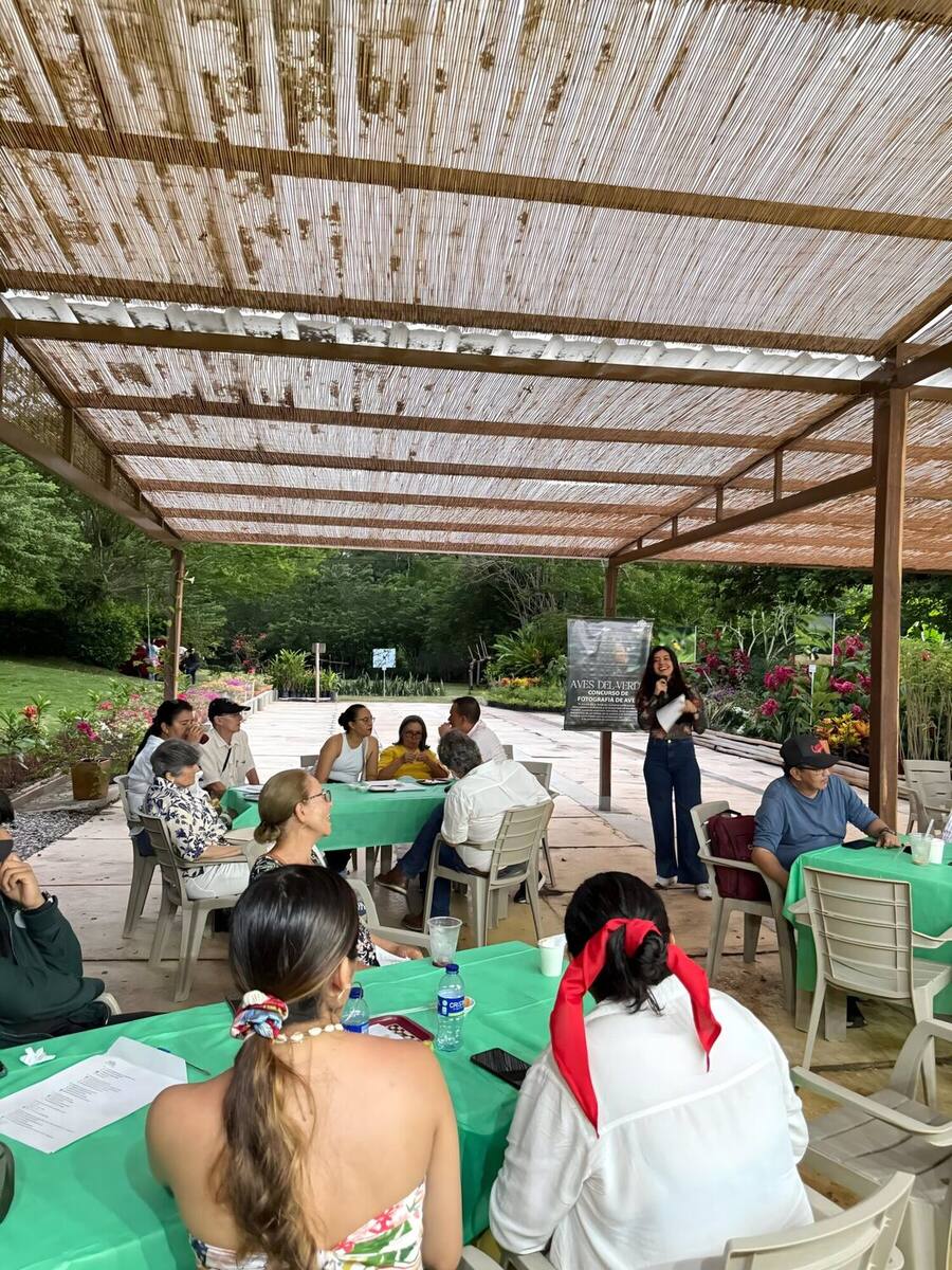 Una mujer con micrófono dirige una charla a un grupo de asistentes sentados en mesas con manteles verdes, bajo la pérgola de caña del parque, durante el concurso de fotografía de aves.