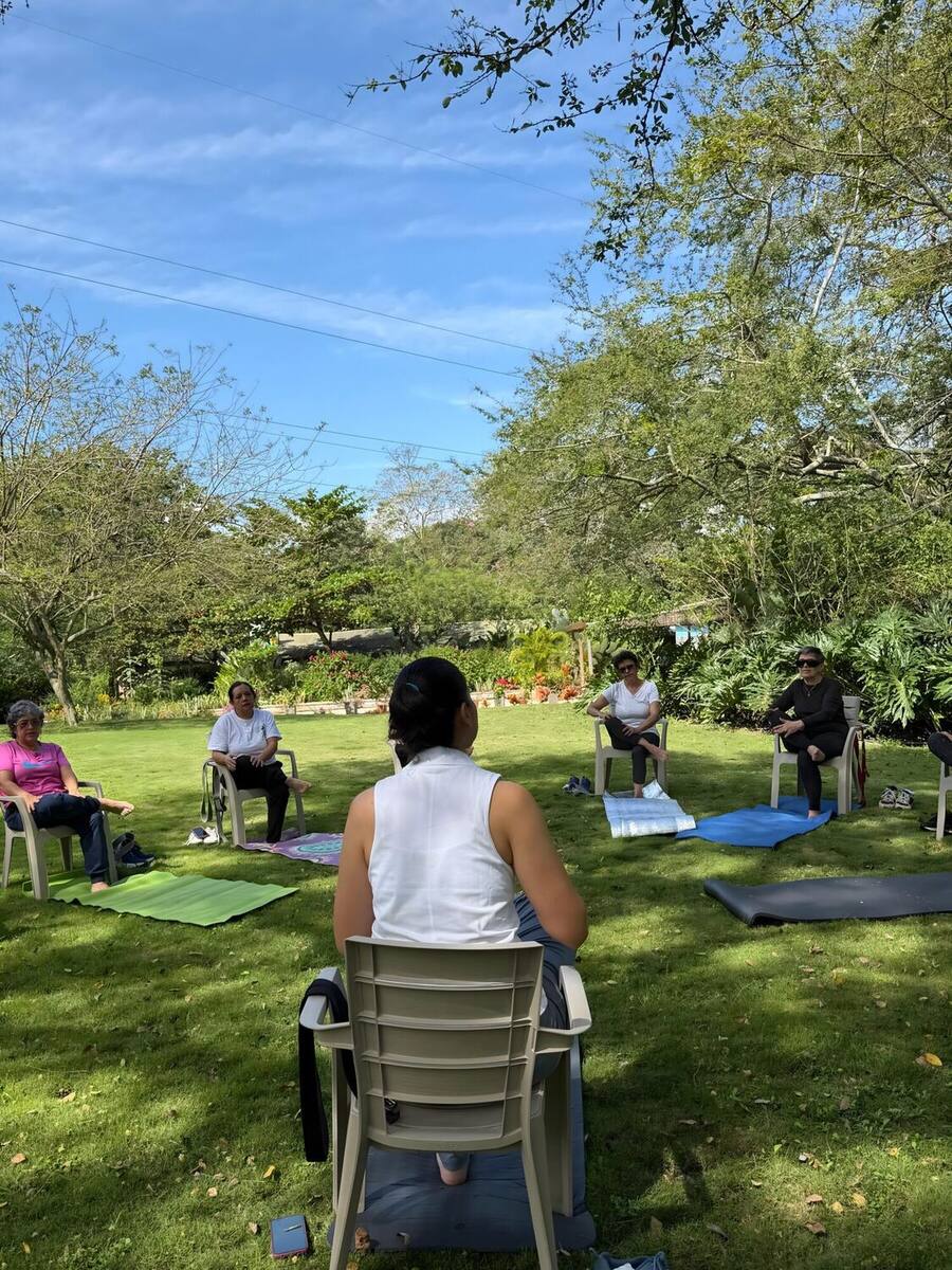 Otra sesión, con la instructora sentada de espaldas guiando al grupo: cinco personas repartidas en sillas sobre el césped, cada una con su esterilla, frente a un macizo de flores naranjas.
