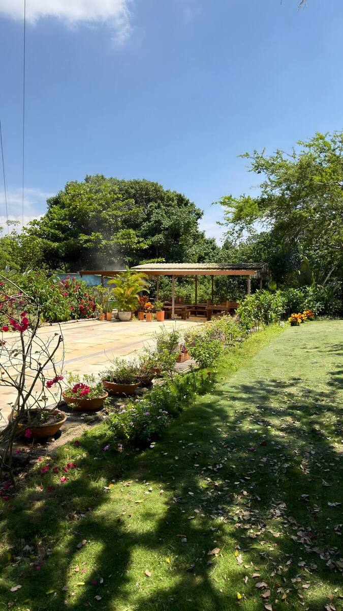 Kiosco de madera con mesas y bancas al fondo de una explanada bordeada por materas de flores y prado.